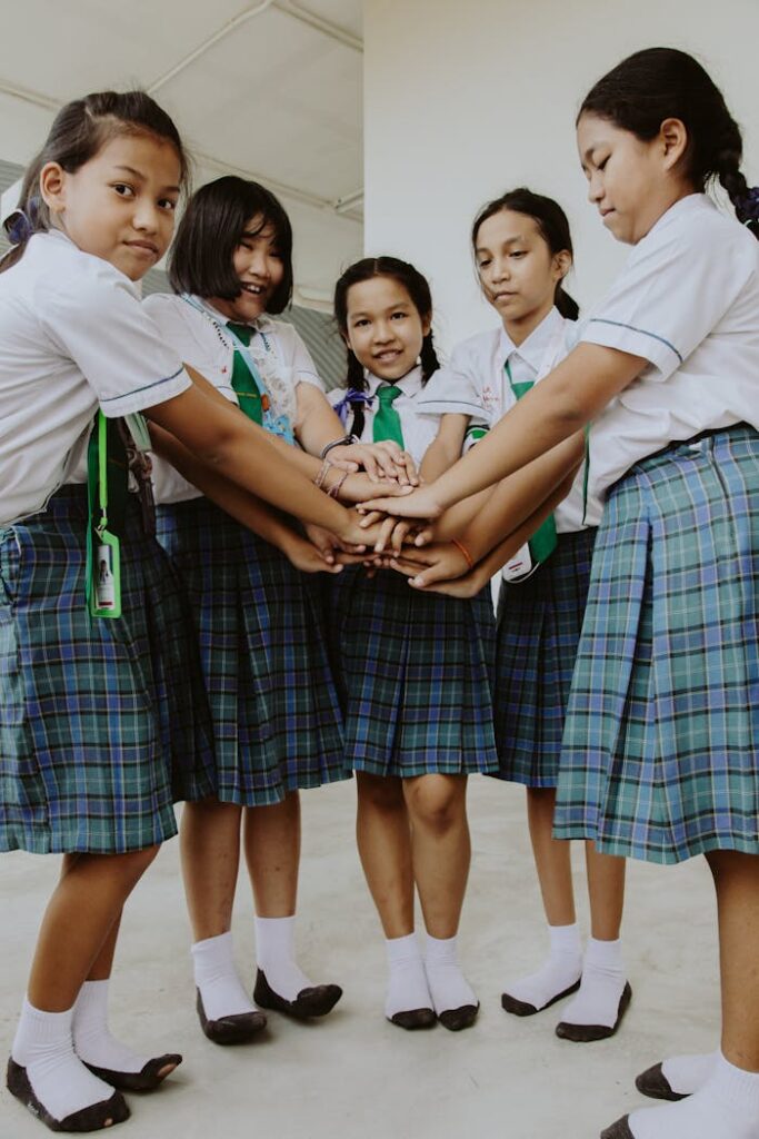 Five schoolgirls in uniform stack hands symbolizing friendship and teamwork indoors.
