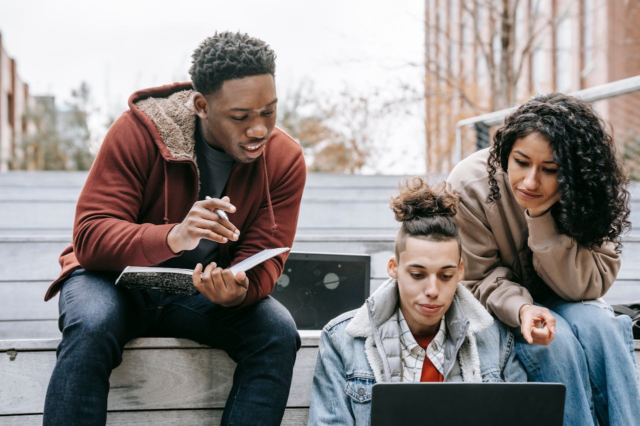 Young multiethnic friends in warm clothes sitting on stairs in campus while preparing for exam and taking notes in notepad with pen and browsing on laptop in daytime