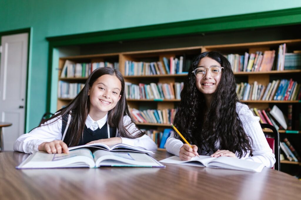 Two cheerful students sit in a library, engaged in study and writing, surrounded by bookshelves.