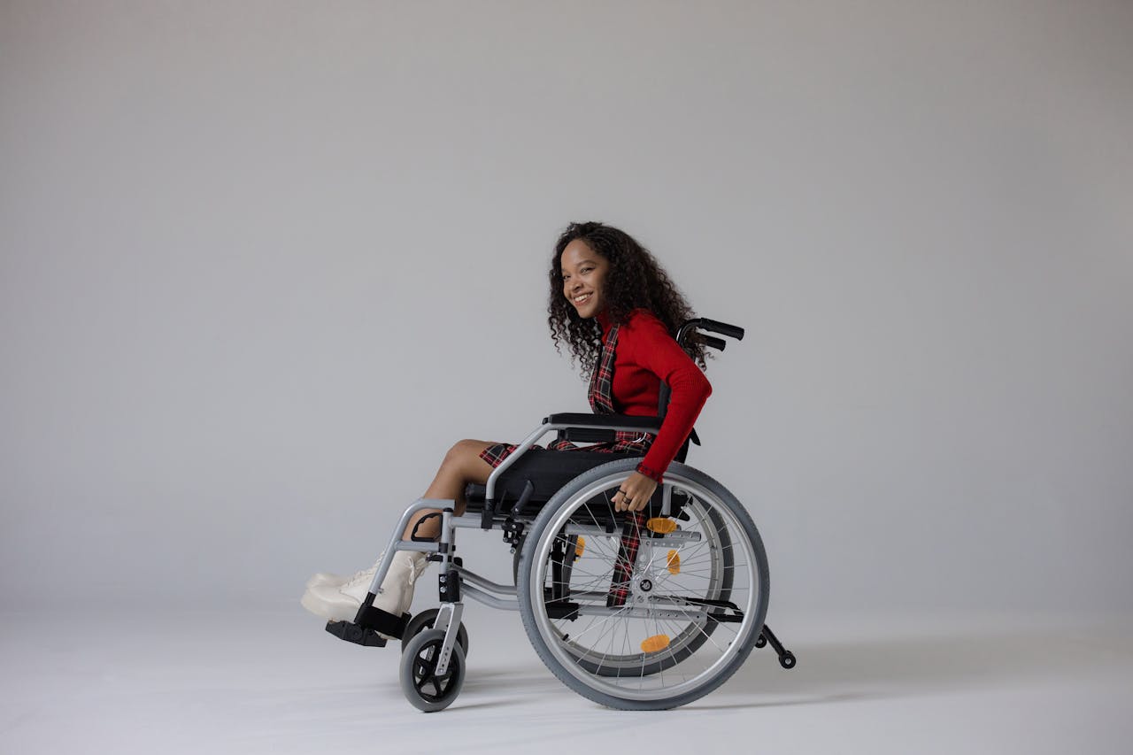A young woman with curly hair smiling while seated in a wheelchair against a gray background.
